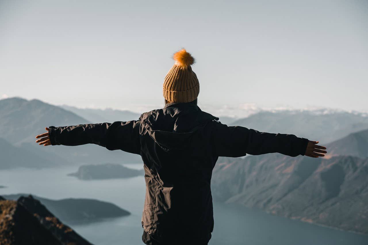 Woman with hands apart against mountain valley Back view of woman in hat and coat standing on high peak holding hands apart against majestic terrain with lake and mountains