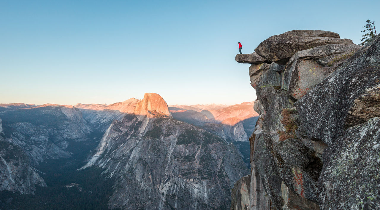 A fearless hiker is standing on an overhanging rock enjoying the view towards famous Half Dome at Glacier Point overlook in beautiful evening twilight, Yosemite National Park, California, USA